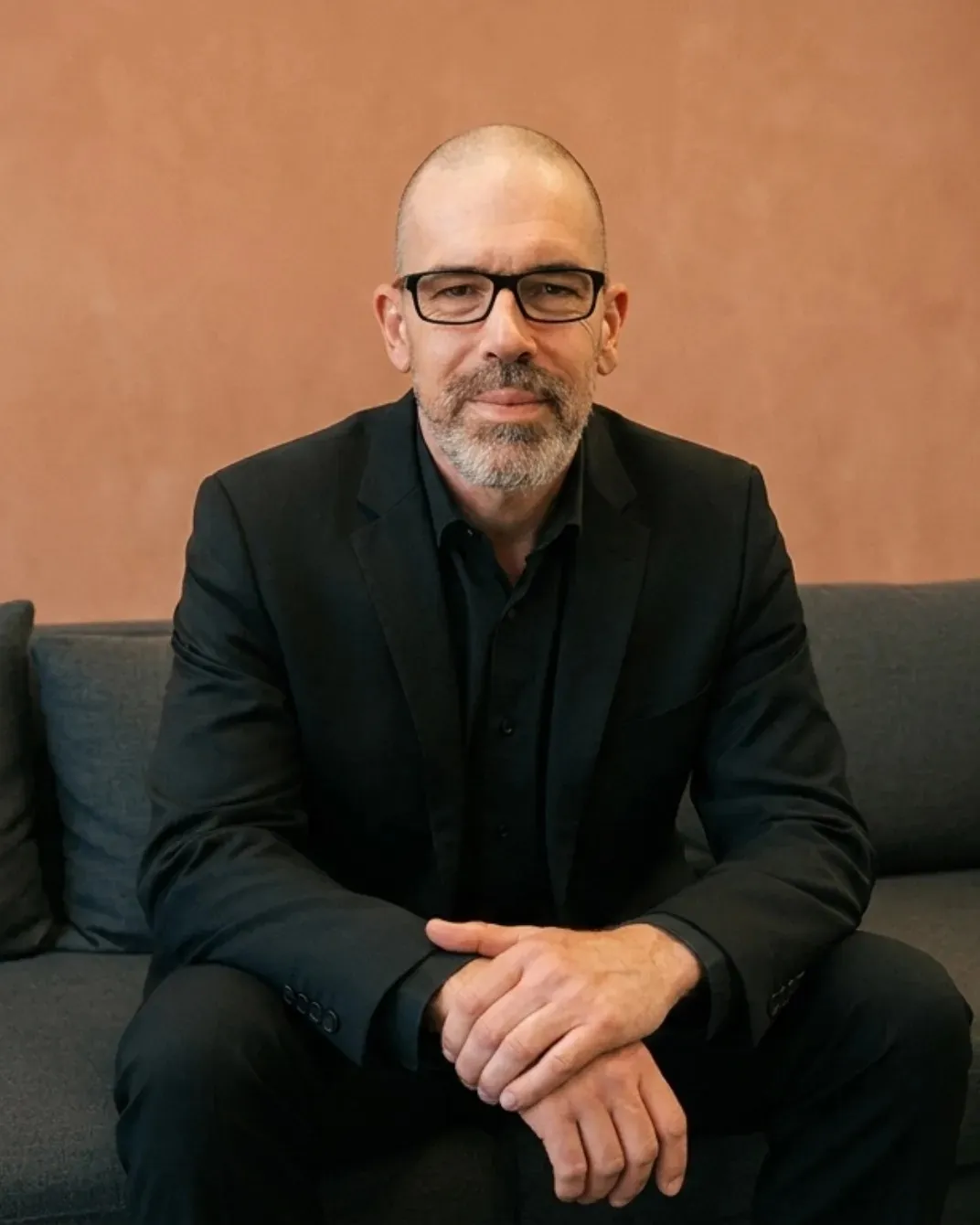 Mark Steven in a black suit and glasses, sitting on a grey couch against a terracotta wall.
